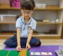 Child in school uniform with glasses sitting on a purple mat in a classroom interacting with Montessori mathematical learning materials Starshine Montessori 90x80