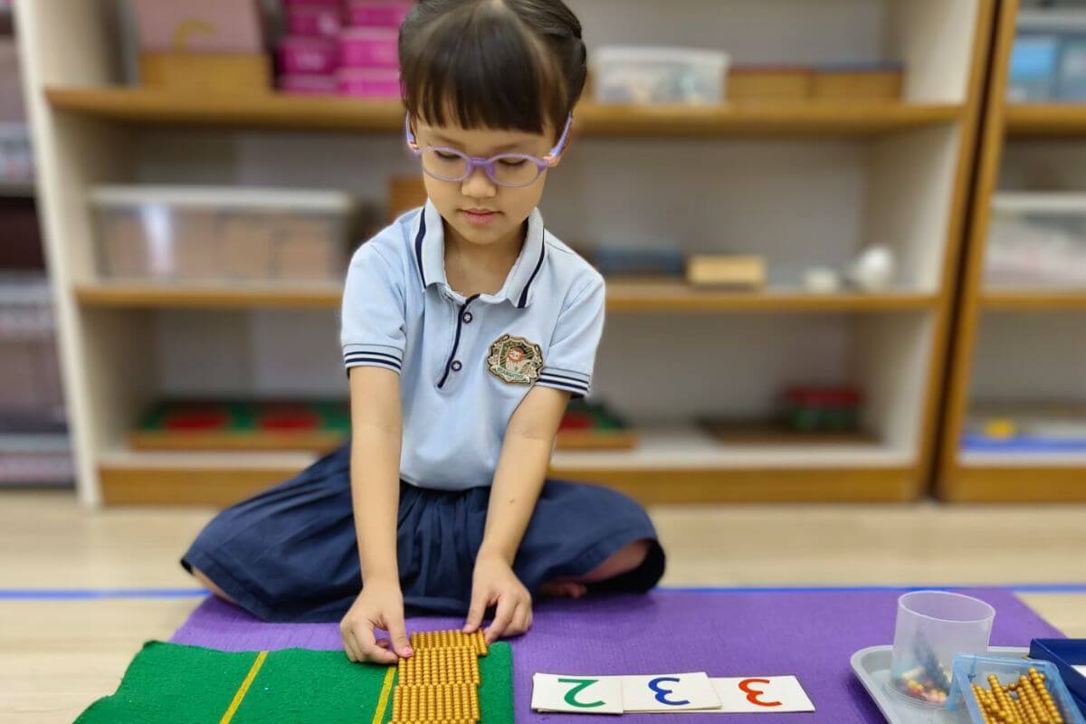 Child in school uniform with glasses sitting on a purple mat in a classroom, interacting with Montessori mathematical learning materials
