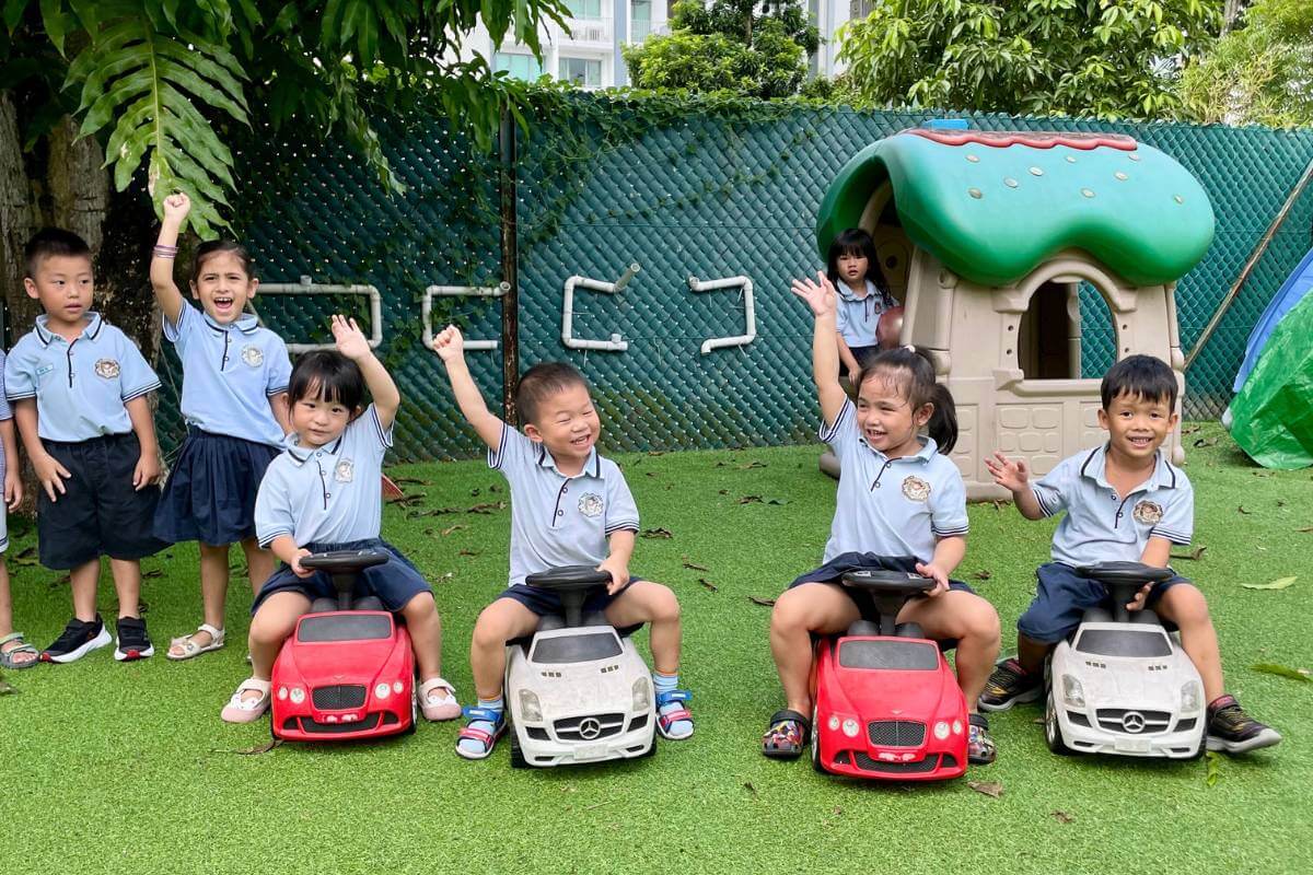 Happy children in school uniforms sitting on toy cars in a playground, with excited expressions as they raise their hands - Starshine Montessori