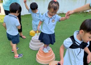 Preschool children in uniforms participating in a balance exercise outdoors, stepping on and off inverted, colored bowls on a grassy surface, with teacher guiding them - Starshine Montessori