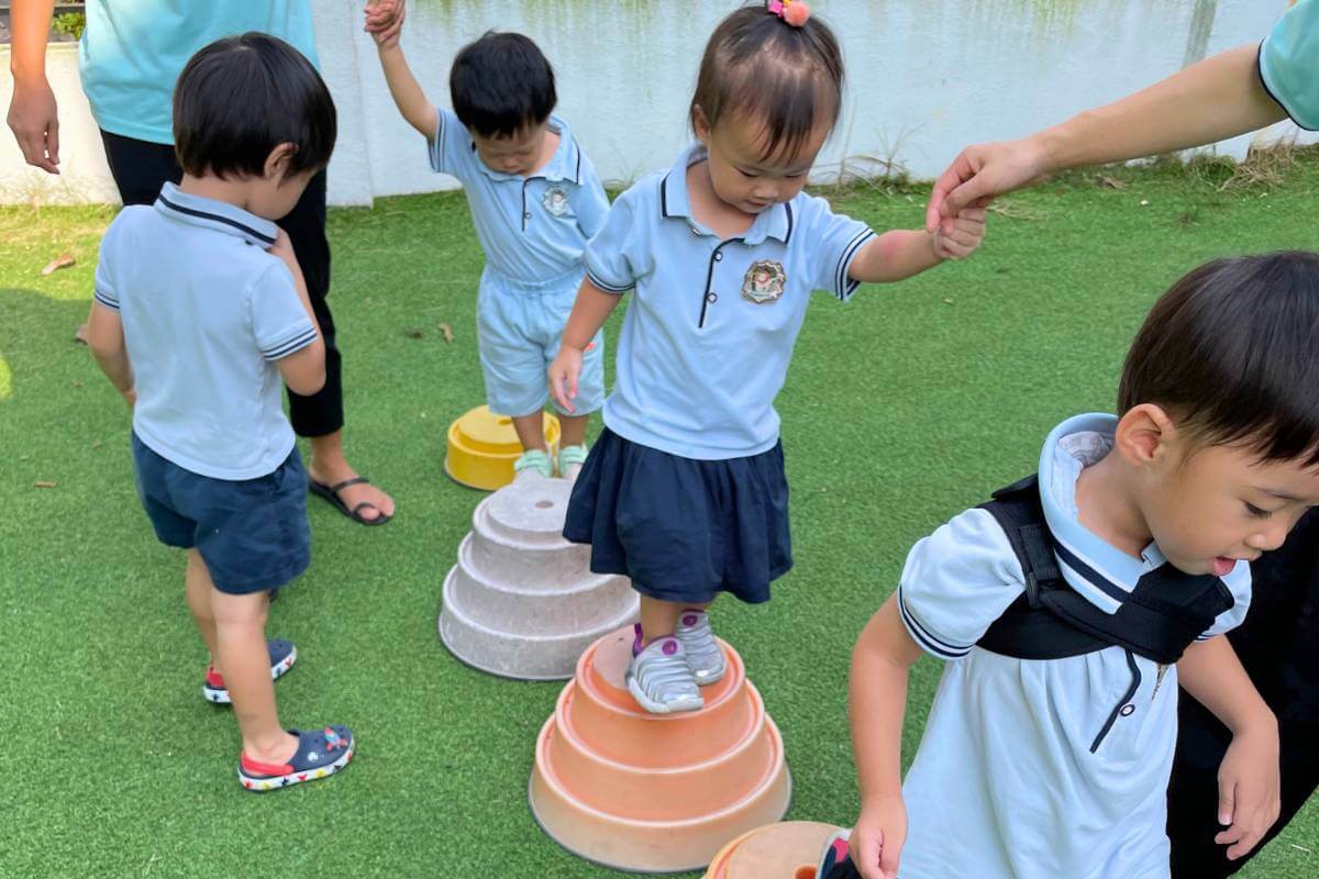 Preschool children in uniforms participating in a balance exercise outdoors, stepping on and off inverted, colored bowls on a grassy surface, with teacher guiding them - Starshine Montessori