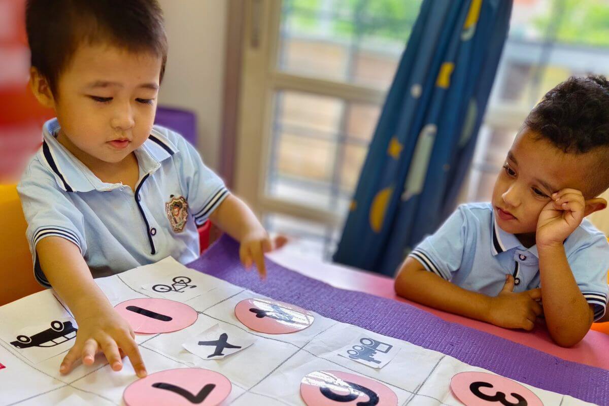 Two children in uniforms at a table with a learning sheet of numbers and symbols, one pointing and the other watching intently