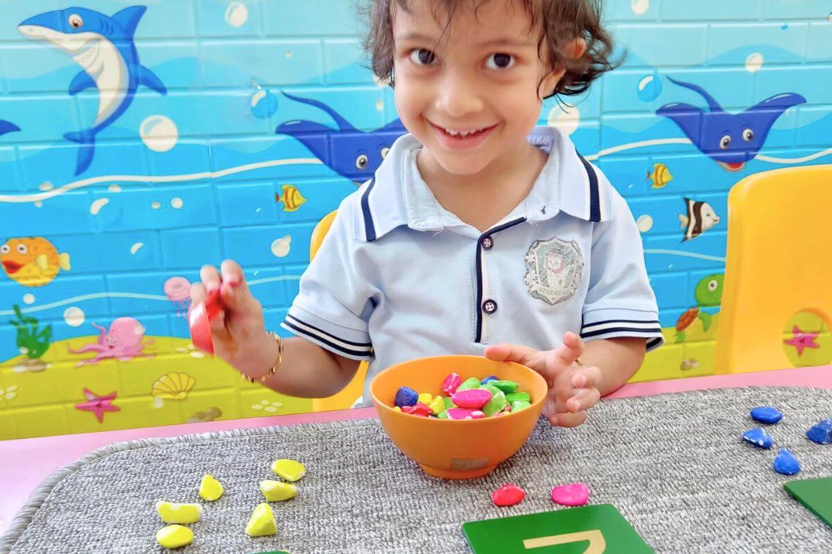 A smiling child engaging in a color matching activity with colorful stones and a bowl, seated in a vibrant classroom with an underwater-themed wall at Starshine Montessori.