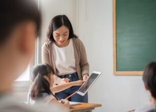 Teacher guiding a student with a tablet in a classroom, using technology for interactive learning and enhancing digital skills.