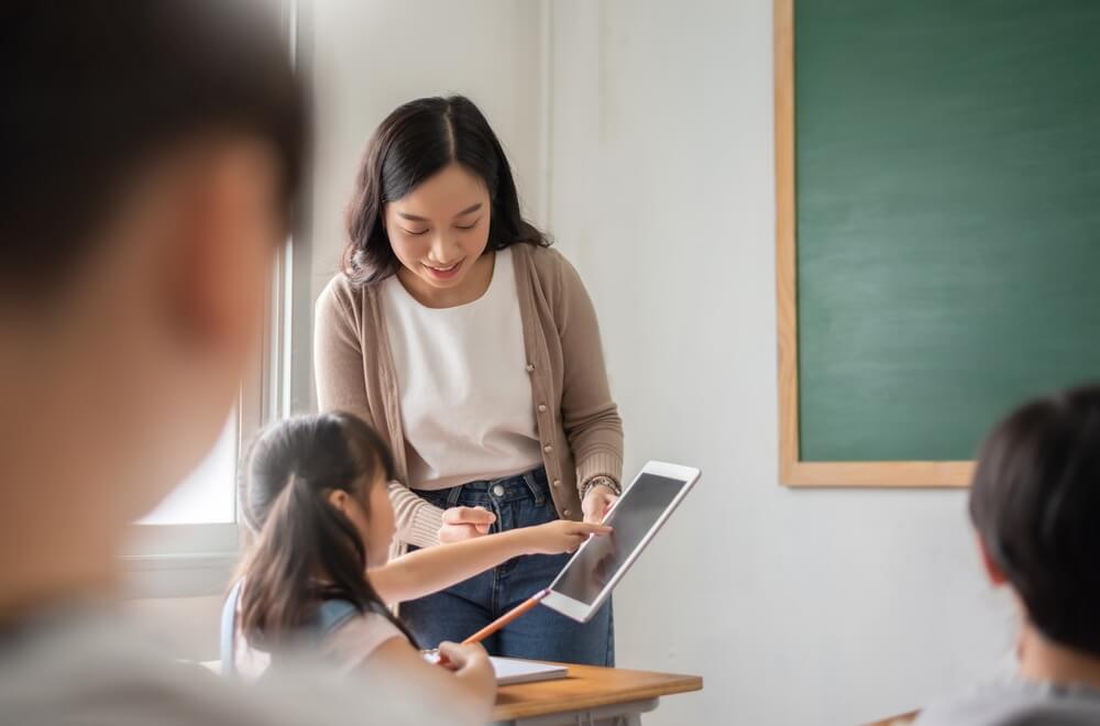 Teacher guiding a student with a tablet in a classroom, using technology for interactive learning and enhancing digital skills.