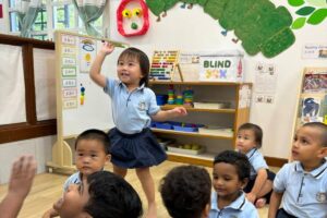 A joyful preschool classroom setting with a child standing and raising her hand enthusiastically among her peers, surrounded by Montessori learning materials and colorful decorations at Starshine Montessori.