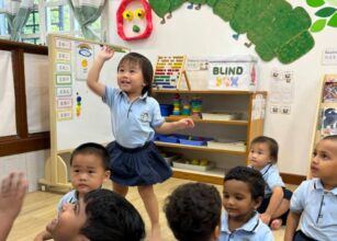 A joyful preschool classroom setting with a child standing and raising her hand enthusiastically among her peers, surrounded by Montessori learning materials and colorful decorations at Starshine Montessori.