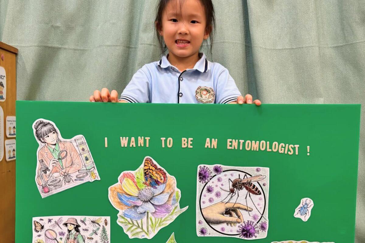 A young girl in a light blue school uniform holding a green presentation board titled "I WANT TO BE AN ENTOMOLOGIST!" with drawings and illustrations of insects, flowers, and entomological tools at Starshine Montessori.