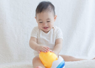 A cheerful baby in a white onesie sitting on a soft white surface, playing with a colorful toy, showcasing moments of joy and exploration in early childhood.