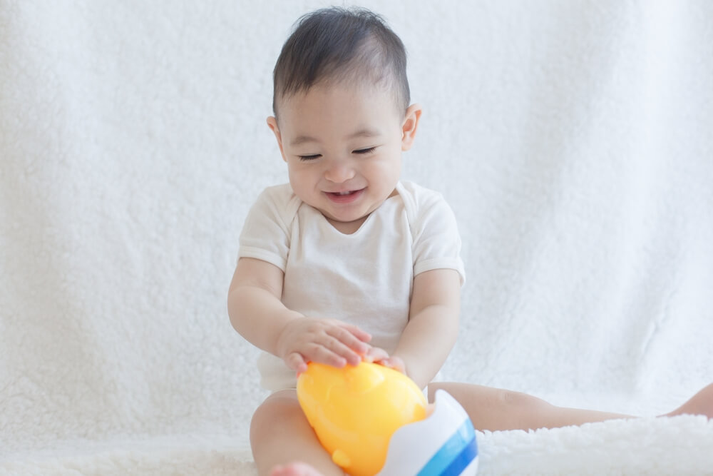 A cheerful baby in a white onesie sitting on a soft white surface, playing with a colorful toy, showcasing moments of joy and exploration in early childhood.