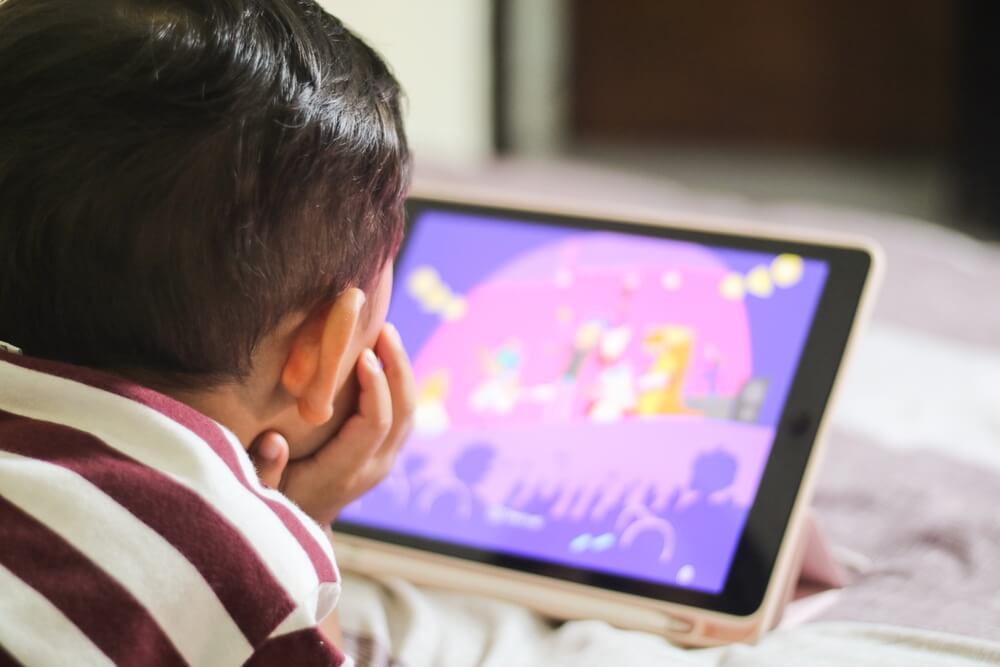 A young child wearing a striped shirt intently watching an animated video on a tablet screen, placed on a stand. The bright purple and pink visuals from the tablet highlight the scene, emphasising digital learning or entertainment for children.