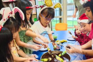 A group of young children engaging in a fun cooking activity, dipping skewered fruits into chocolate, while wearing festive bunny ears and Santa hats. The scene highlights collaboration, creativity, and celebration in a classroom setting at Starshine Montessori.