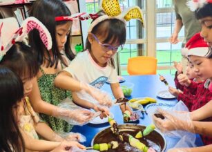 A group of young children engaging in a fun cooking activity, dipping skewered fruits into chocolate, while wearing festive bunny ears and Santa hats. The scene highlights collaboration, creativity, and celebration in a classroom setting at Starshine Montessori.