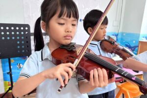 A young girl attentively playing the violin during a music lesson, demonstrating focus and precision. A boy in the background is also playing the violin, emphasising collaborative learning in a classroom at Starshine Montessori.