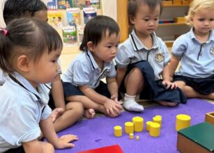 A group of young children sitting attentively in a classroom, exploring yellow wooden cylinder blocks arranged on a purple mat at Starshine Montessori.