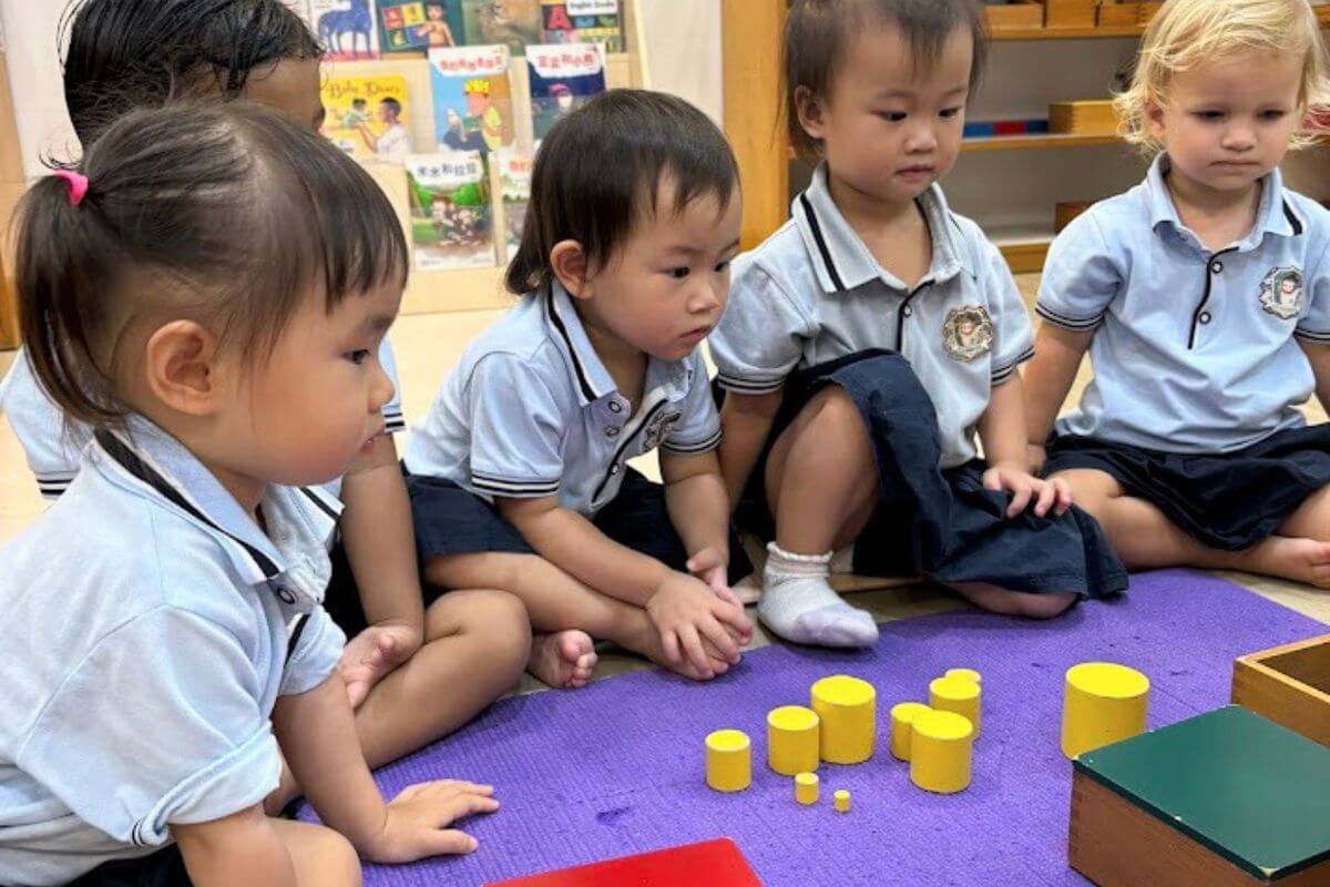 A group of young children sitting attentively in a classroom, exploring yellow wooden cylinder blocks arranged on a purple mat at Starshine Montessori.