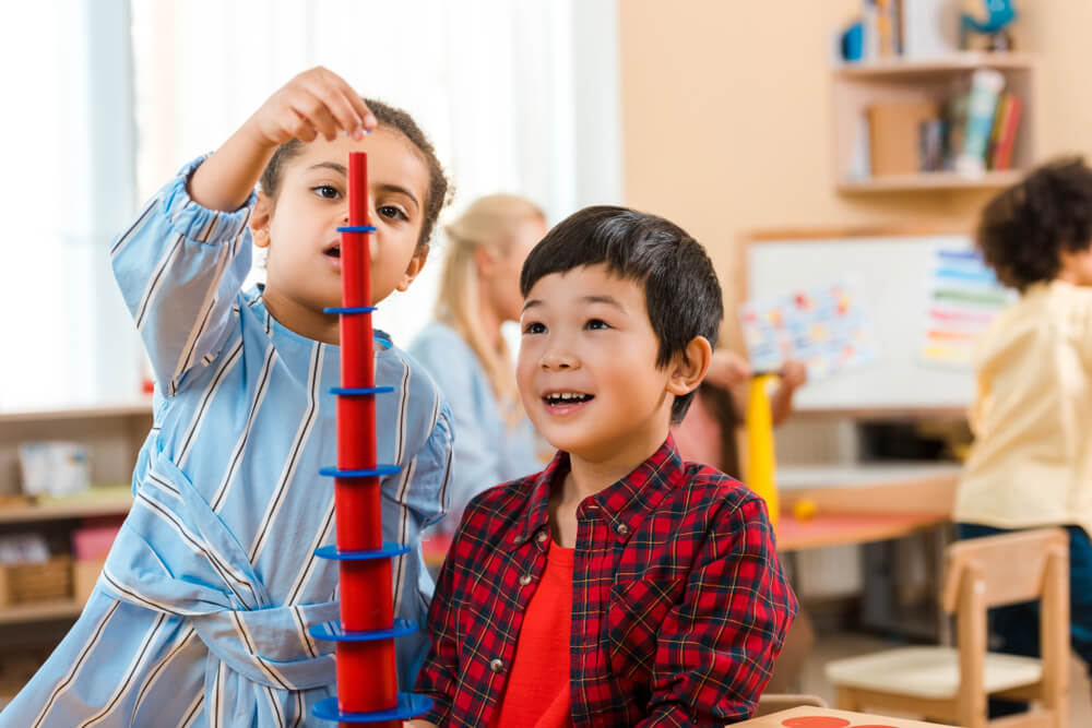 Two young children in a classroom building a tall tower with red and blue blocks, showcasing teamwork and concentration.