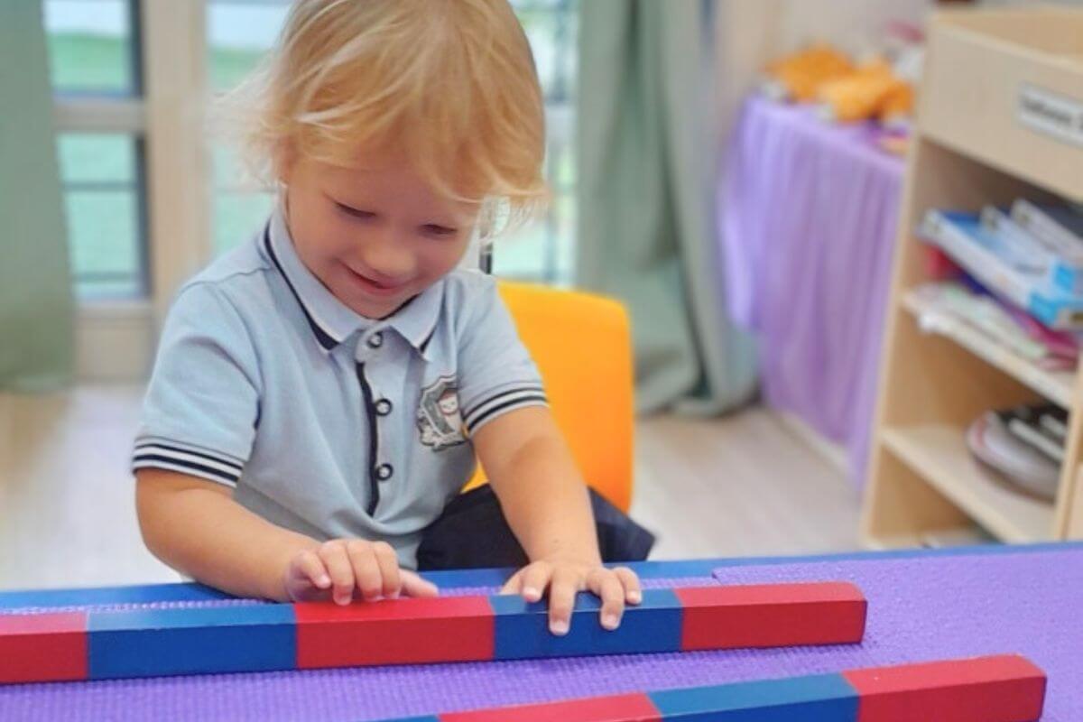 A smiling young child engaging in a Montessori activity with red and blue color blocks arranged on a purple mat in a classroom setting at Starshine Montessori.