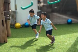 Two young children in matching uniforms running and playing joyfully in an outdoor playground with swings and artificial grass at Starshine Montessori. A fun and engaging Montessori-inspired learning environment promoting physical activity and social development.