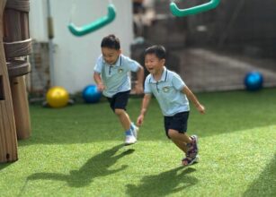 Two young children in matching uniforms running and playing joyfully in an outdoor playground with swings and artificial grass at Starshine Montessori. A fun and engaging Montessori-inspired learning environment promoting physical activity and social development.