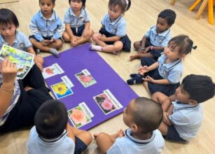 A Montessori teacher is conducting a botany lesson with young children sitting in a circle on the floor. The teacher holds a picture card while the children attentively observe various labeled flower cards arranged on a purple mat at Starshine Montessori.