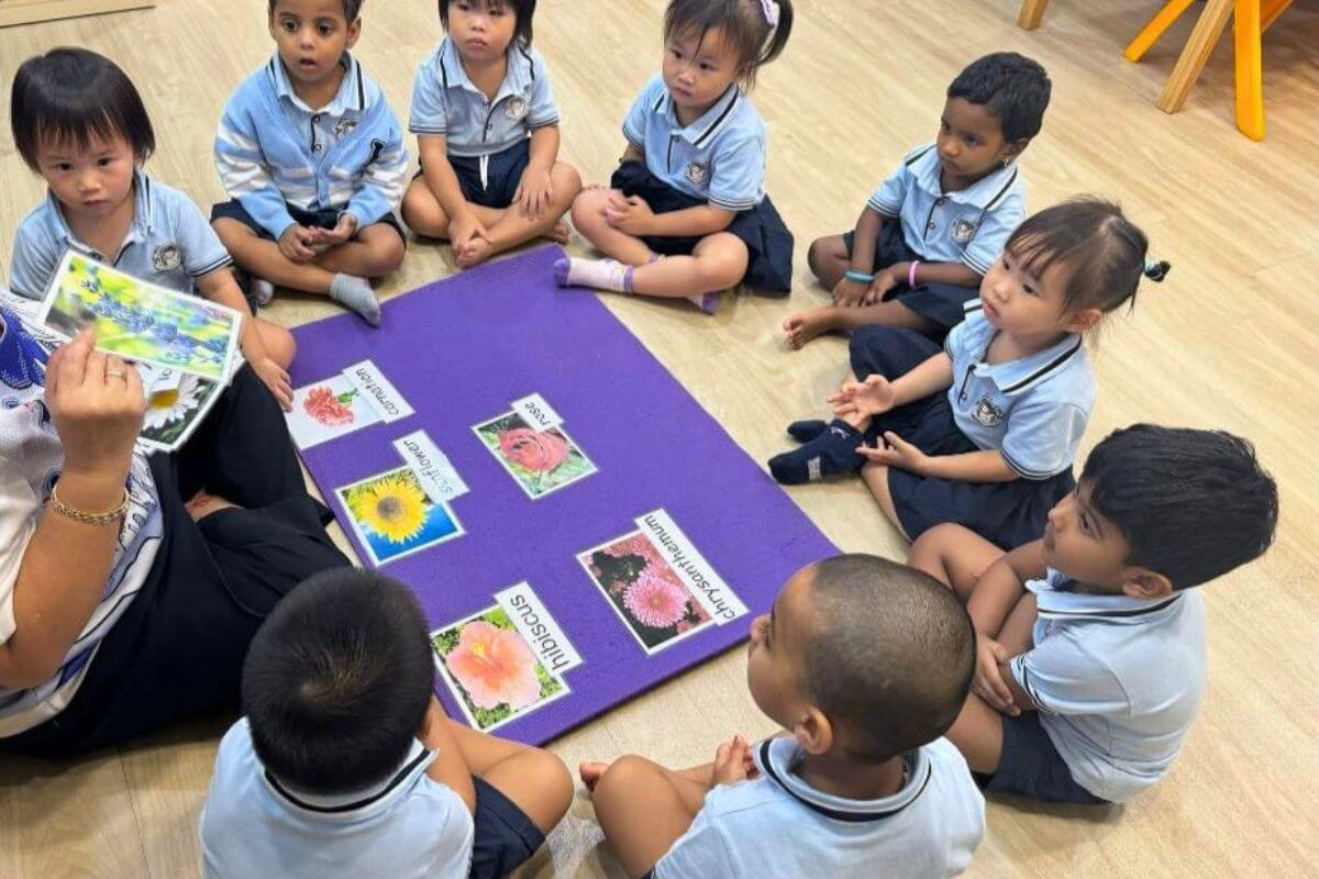 A Montessori teacher is conducting a botany lesson with young children sitting in a circle on the floor. The teacher holds a picture card while the children attentively observe various labeled flower cards arranged on a purple mat at Starshine Montessori.