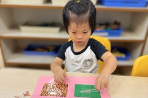 A young child in a Montessori classroom is engaged in a texture matching activity. The child is carefully placing a transparent card on a pink sensory board featuring different textured and patterned squares, reinforcing tactile learning and sensory exploration at Starshine Montessori.