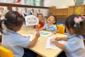 Young children engaged in a fun coloring activity in a Montessori classroom at Starshine Montessori. They are seated around a table, wearing uniforms, and expressing joy while working on a school bus-themed worksheet. The learning environment features educational materials, personalized student portraits, and Montessori-inspired tools for early childhood education.