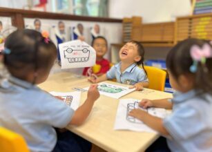 Young children engaged in a fun coloring activity in a Montessori classroom at Starshine Montessori preschool. They are seated around a table, wearing uniforms, and expressing joy while working on a school bus-themed worksheet. The learning environment features educational materials, personalized student portraits, and Montessori-inspired tools for early childhood education.