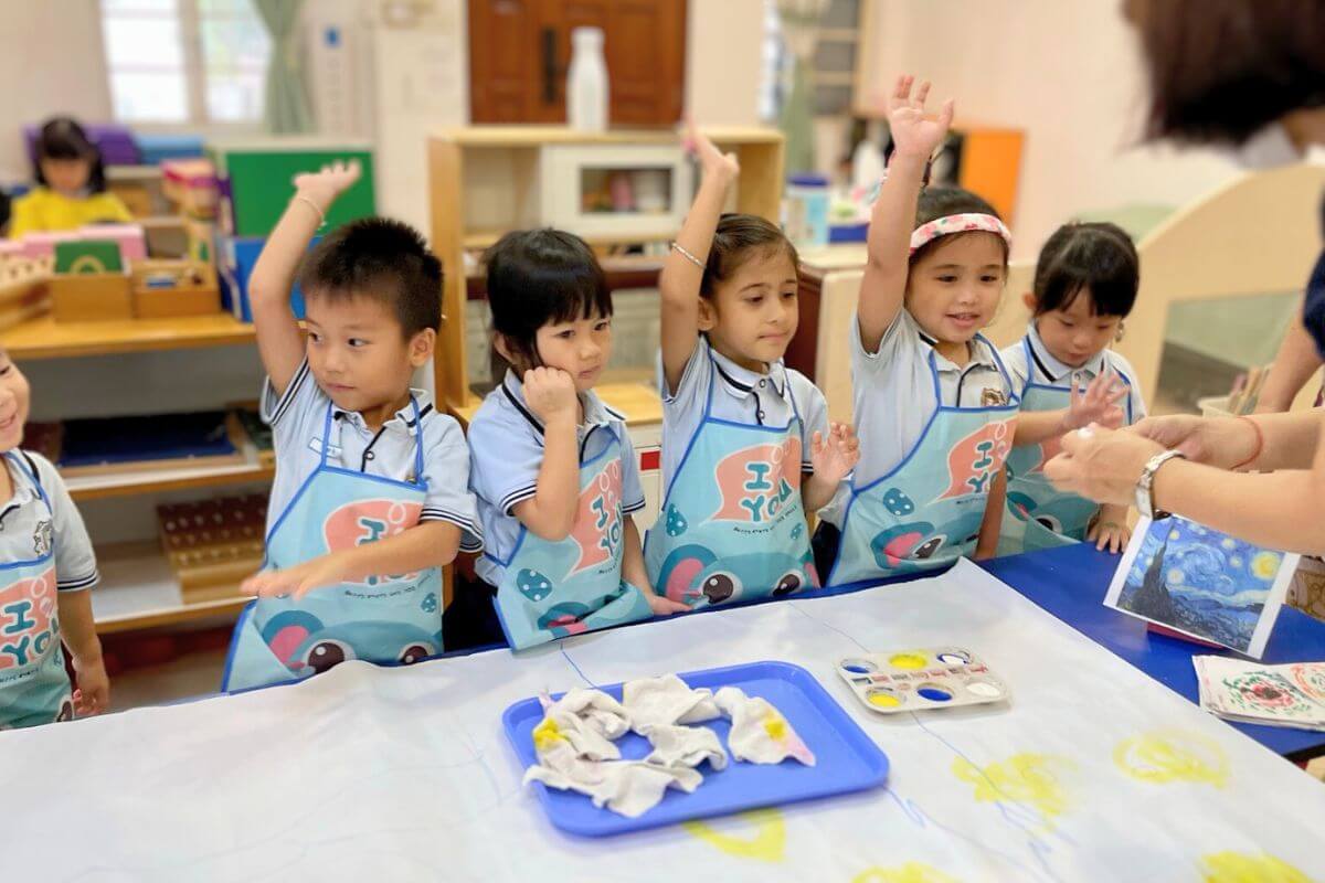 Young children in a Montessori classroom at Starshine Montessori preschool participating in an interactive art painting activity, wearing aprons and engaging with their teacher. The learning environment encourages creativity, self-expression, and hands-on exploration through guided artistic experiences.