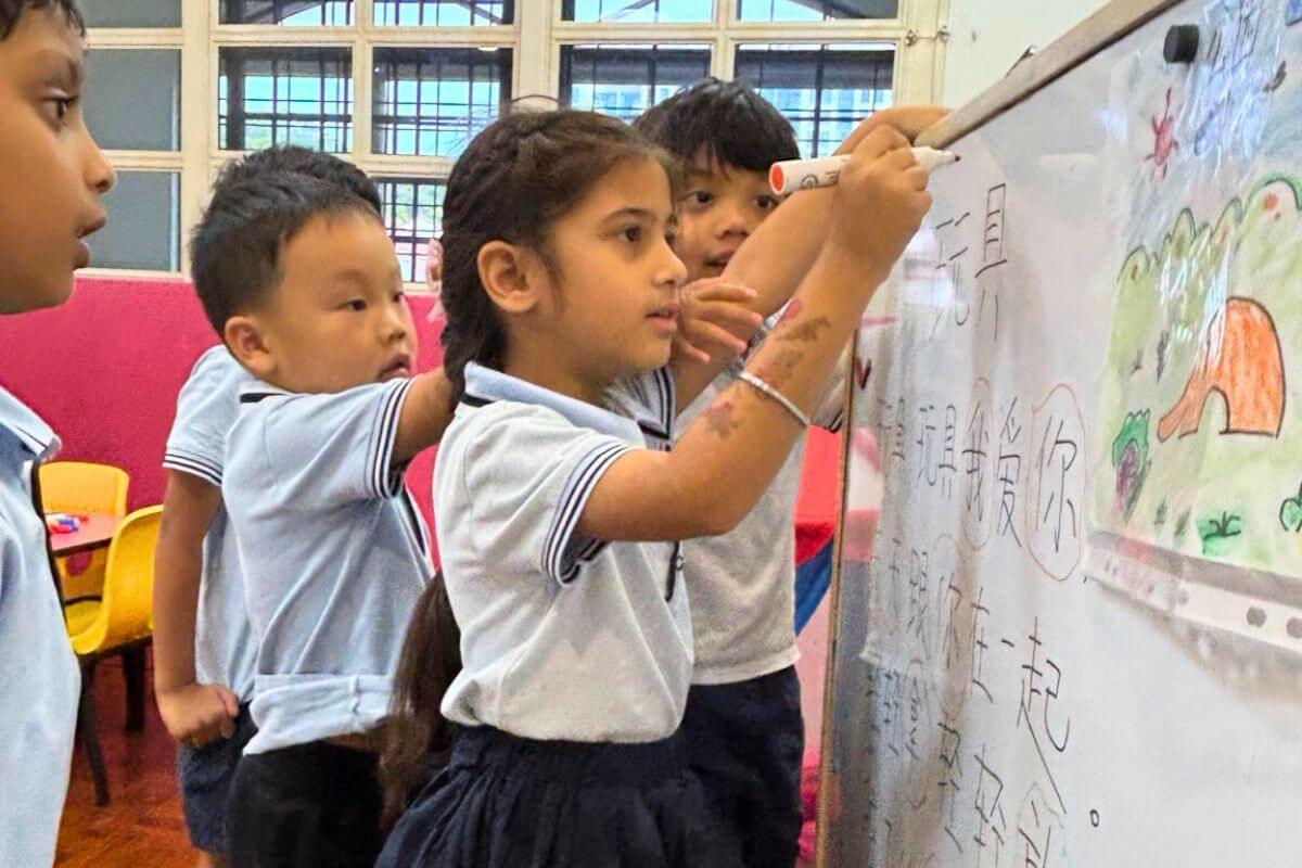 Group of young children participating in a collaborative whiteboard activity in a Montessori classroom at Starshine Montessori Preschool. The students are writing and drawing on the board, practicing language skills and creative expression, fostering teamwork and hands-on learning.